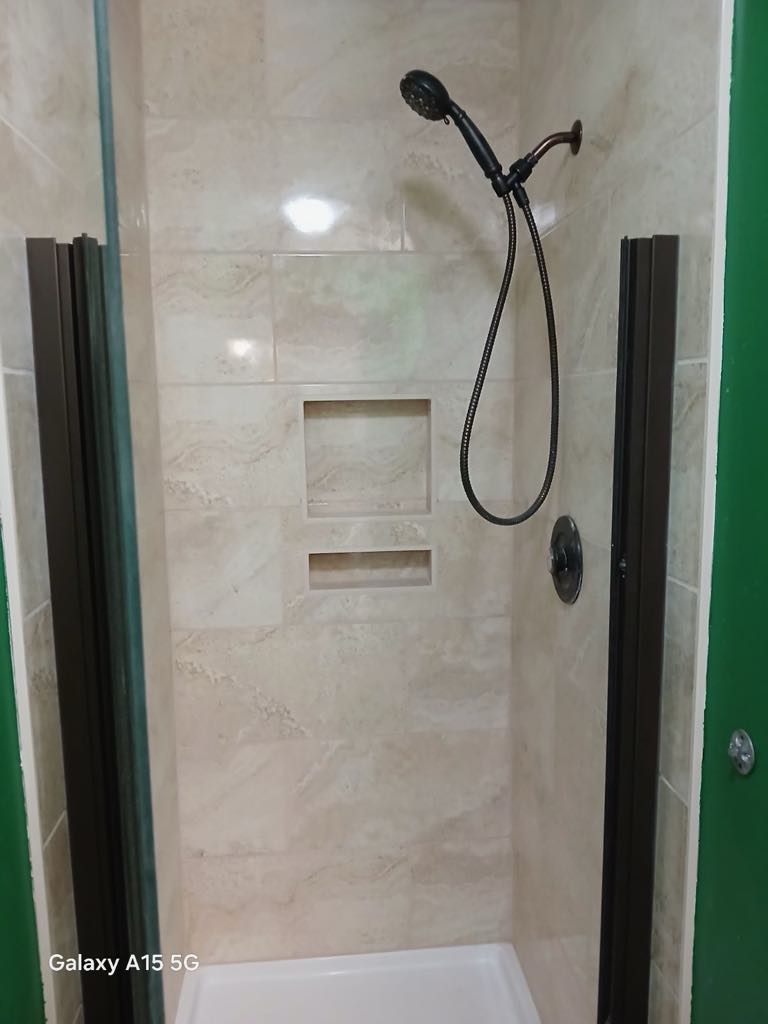 A small, tiled shower with a rain showerhead, a glass door, and two built-in wall niches for storage offers a perfect example of modern bathroom remodeling. The light beige tiles complement the design, creating a clean and contemporary look.