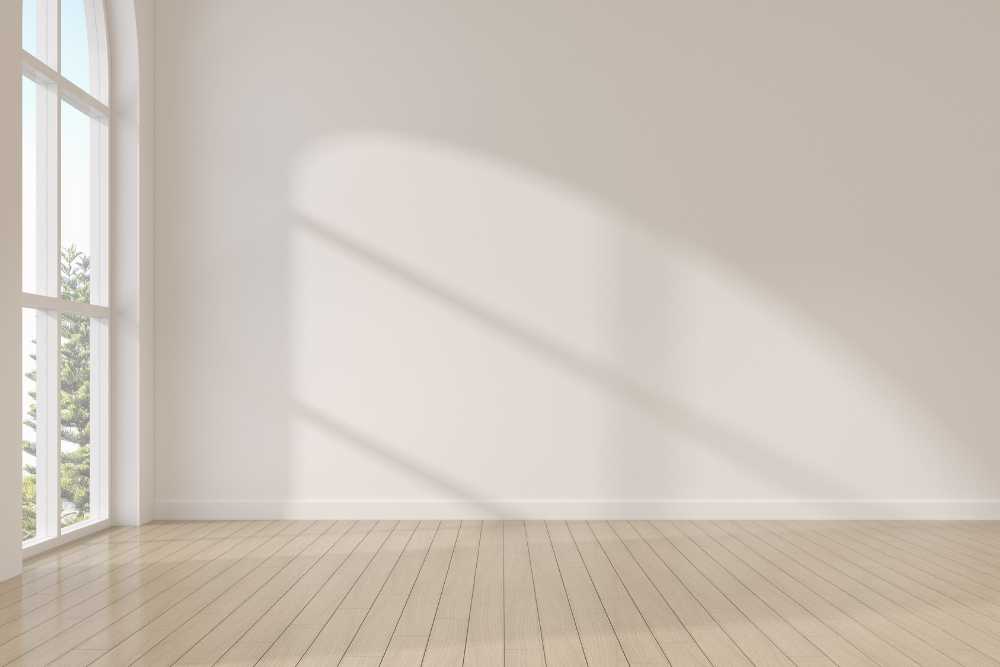 A bright, empty room with light wooden floors and a large arched window hints at potential kitchen remodeling. Sunlight streams in, casting shadows on the plain white walls. A glimpse of trees is visible outside, adding a touch of nature to this inviting space.