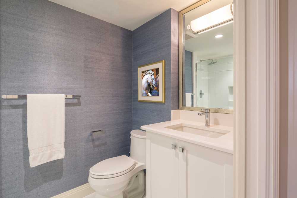 A modern bathroom with blue textured walls showcases expert bathroom remodeling, featuring a white vanity and sink, a wall-mounted mirror, a toilet, and a towel holder with a white towel. Above the toilet hangs a framed picture of a horse.