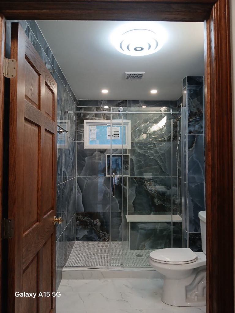 A modern bathroom featuring dark marble tiles and a glass-enclosed shower invites relaxation after a successful bathroom remodeling project. The space is complete with a white toilet, wooden door, recessed lighting, and a ceiling vent.