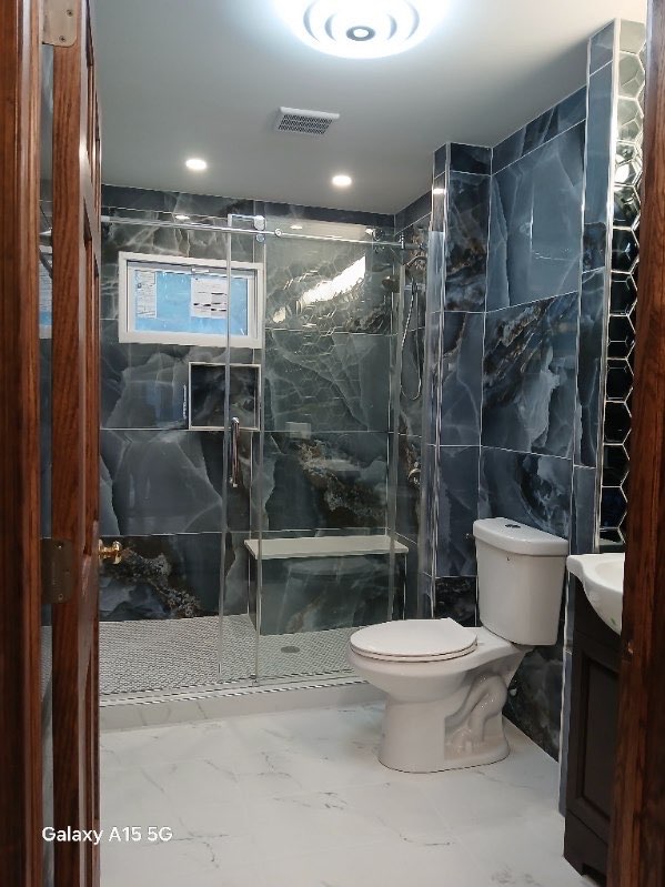 A modern bathroom showcasing dark marble walls, a glass-enclosed shower with a built-in bench, and a white toilet. The light-colored marble floor contrasts with the dark wooden door. Ceiling lights illuminate the space, making it a perfect example of sleek bathroom remodeling.