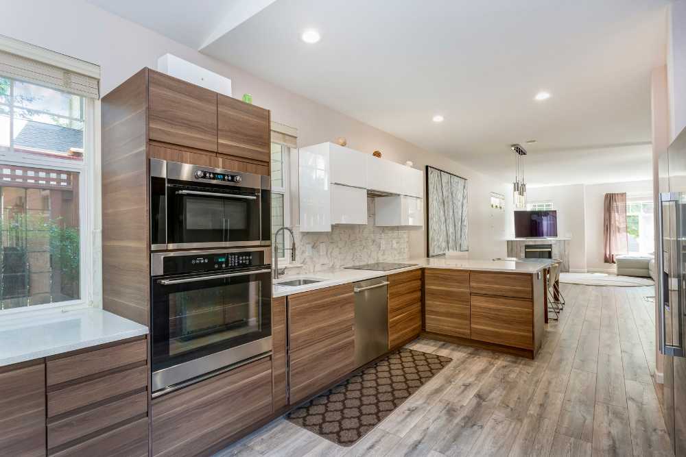 A modern kitchen with wooden cabinets, two built-in ovens, a dishwasher, and a marble backsplash. The space, ideal for interior painting color schemes, boasts a large island with a light countertop. It features wooden flooring and recessed lighting, offering a view into the living area.