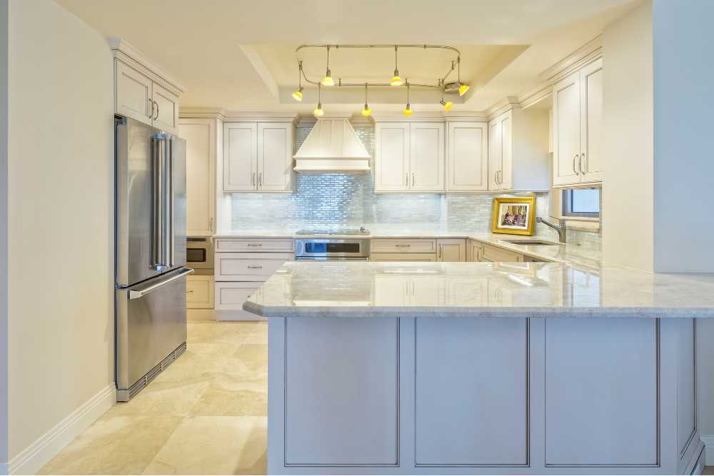 Modern kitchen featuring cream cabinets, stainless steel appliances, and a large central island—perfect for any kitchen remodeling project. A silver refrigerator stands to the left, while a stove with a white range hood is at the back. Overhead lighting illuminates the space.