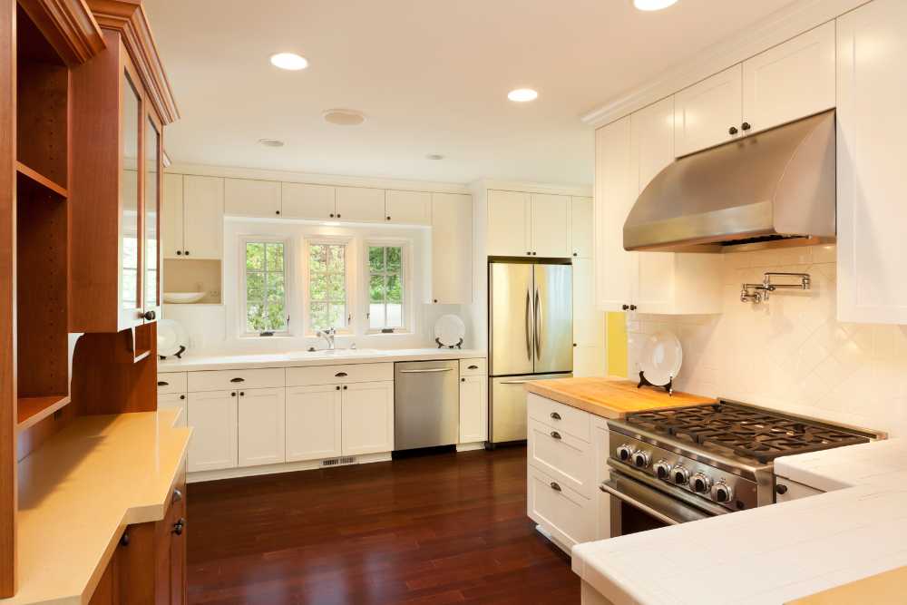A modern kitchen with white cabinets, a large stainless steel oven and range hood, a refrigerator, and a dishwasher. Natural light streams in through a window above the sink. Dark wooden floors contrast the light cabinetry, creating an inviting space perfect for any kitchen remodeling project.
