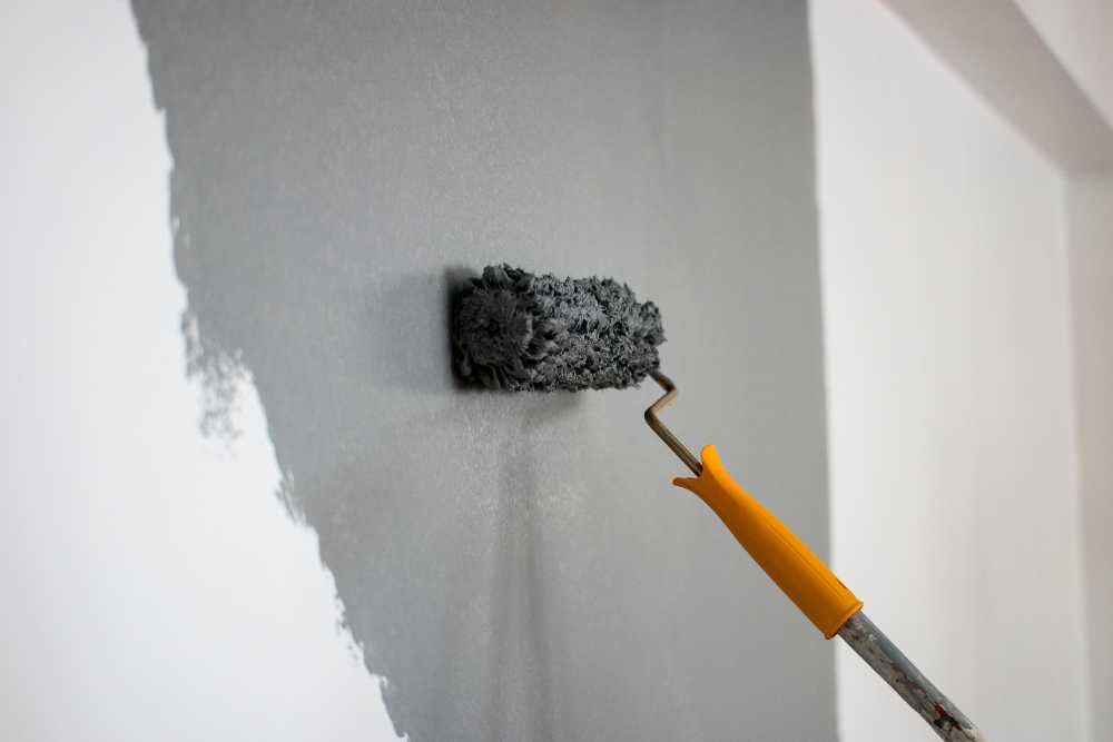 A paint roller with a yellow handle applies gray paint to a white wall, making it perfect for kitchen remodeling by providing a modern, sleek finish.