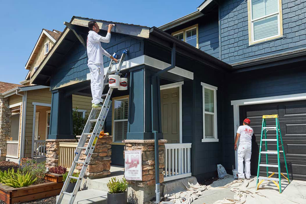 Two workers in white uniforms are meticulously painting the exterior of a two-story house. One stands on a ladder touching up the eaves, while the other focuses near the garage. The navy blue house with white trim gleams against drop cloths covering the ground, ready for interior painting next.