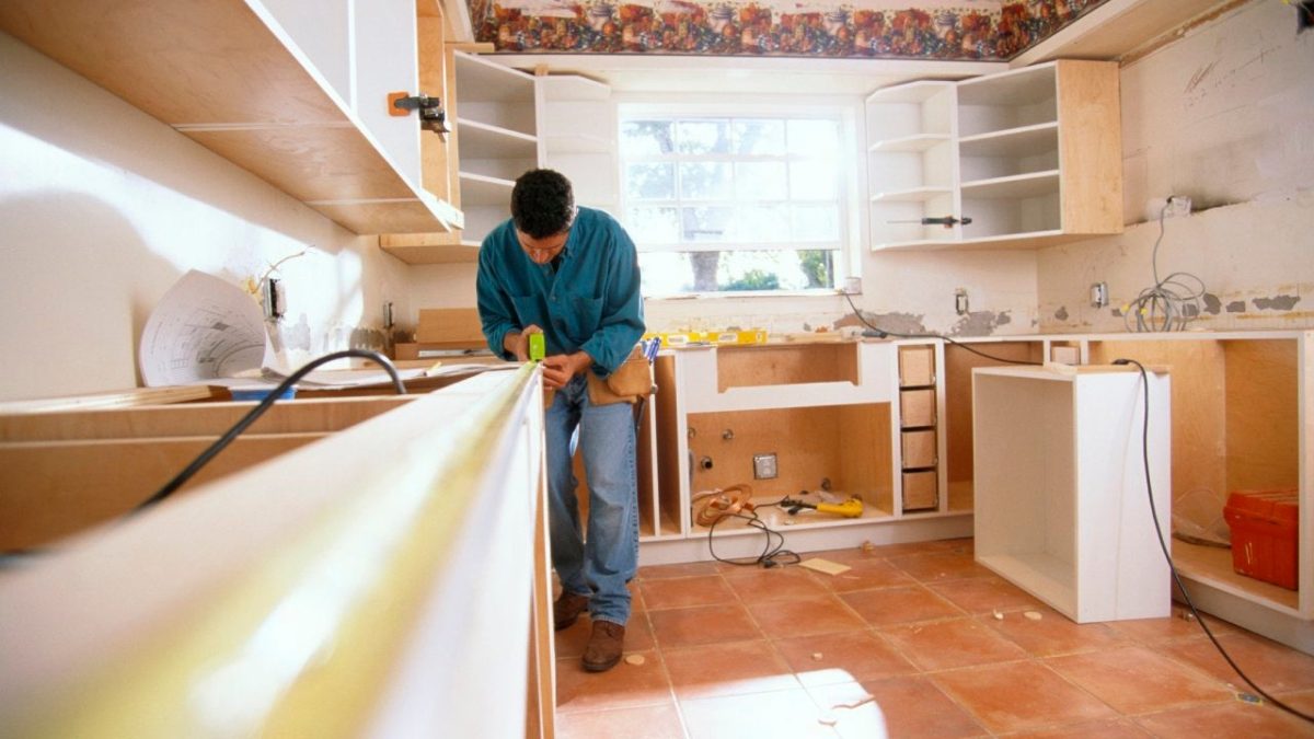 A person measuring a countertop in a kitchen undergoing remodeling. The room features white cabinets, some without doors, and tools scattered on the floor. Exposed walls hint at progress, while natural light filters through a window, illuminating the transformation of this kitchen remodeling project.