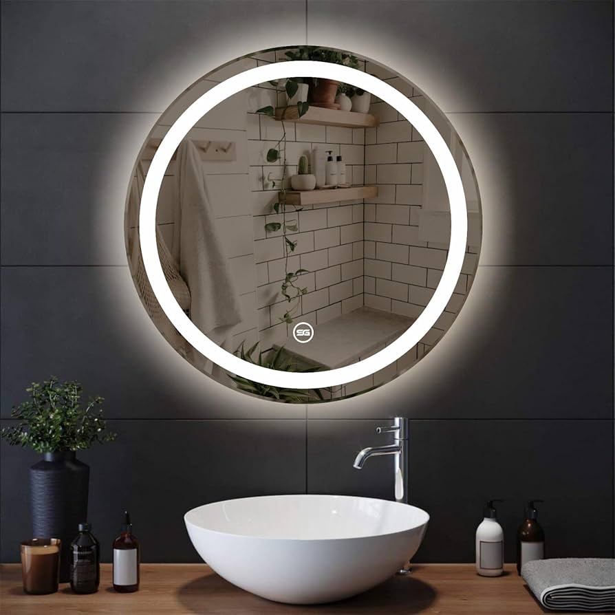 A modern bathroom remodeling showcases a round, backlit mirror reflecting white subway tiles and wooden shelves adorned with plants. Below the mirror, a sleek white vessel sink sits gracefully, surrounded by soap dispensers and a plant on a dark countertop.